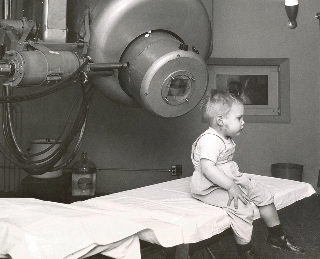 A small patient sits on an exam table, next a linear accelerator device used in 1956 to treat the child's eye tumor.